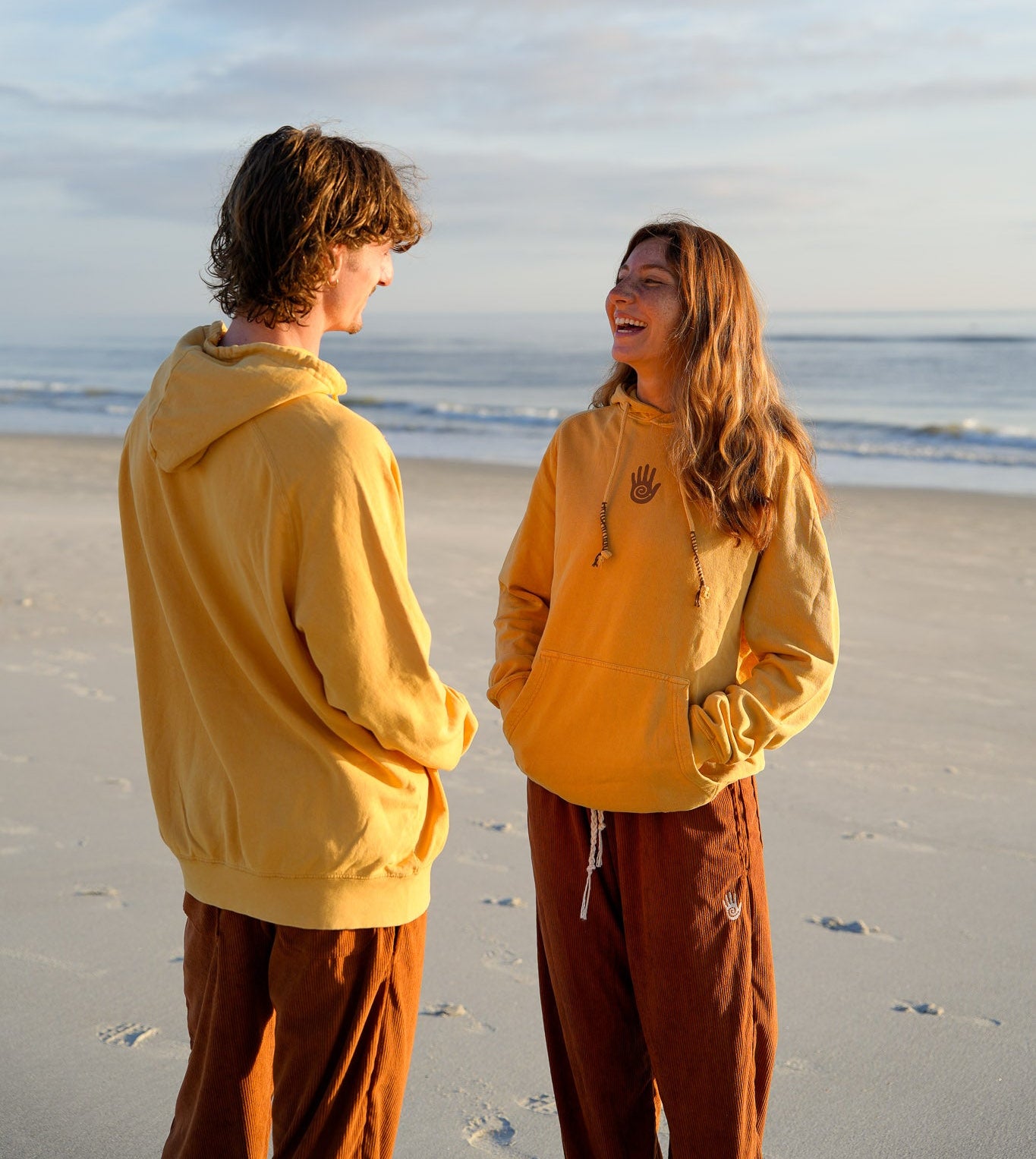 Two people in yellow hoodies standing on a beach with ocean and sky in the background