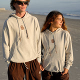 Two people wearing matching light gray hoodies with a hand design on a beach.
