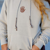 Person wearing a light gray hoodie with a hand design on the chest, standing against a blurred natural background.