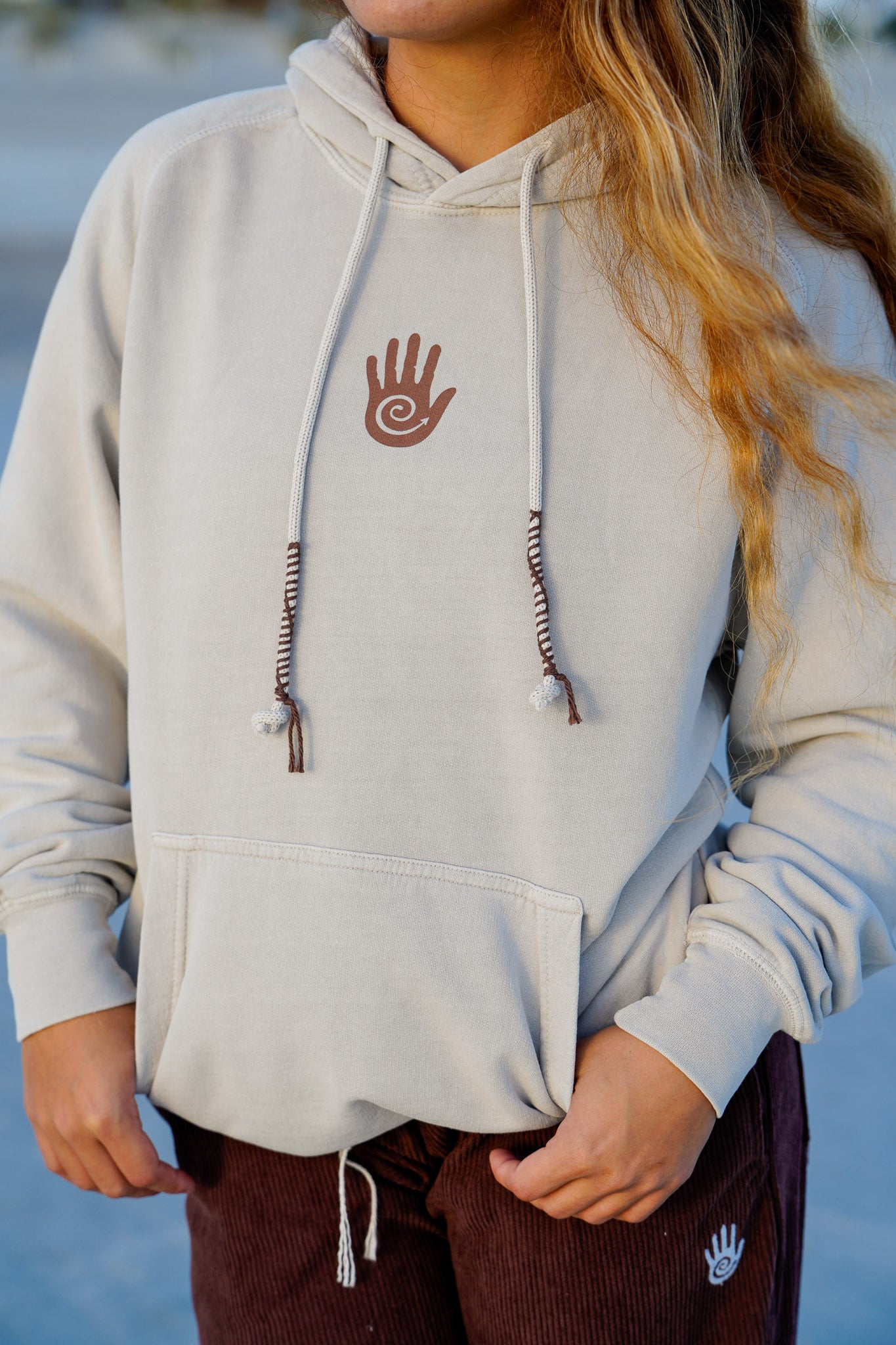 Person wearing a light gray hoodie with a hand design on the chest, standing against a blurred natural background.