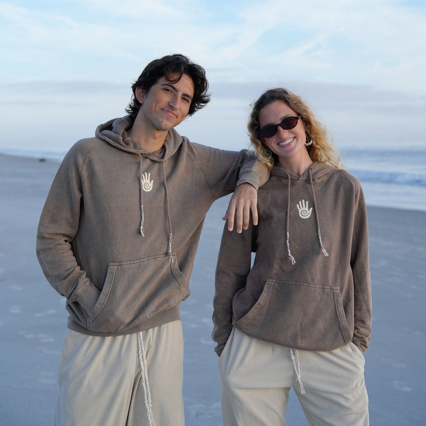 Two people wearing matching brown hoodies on a beach with a clear blue sky.