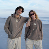 Two people wearing matching brown hoodies on a beach with a clear blue sky.