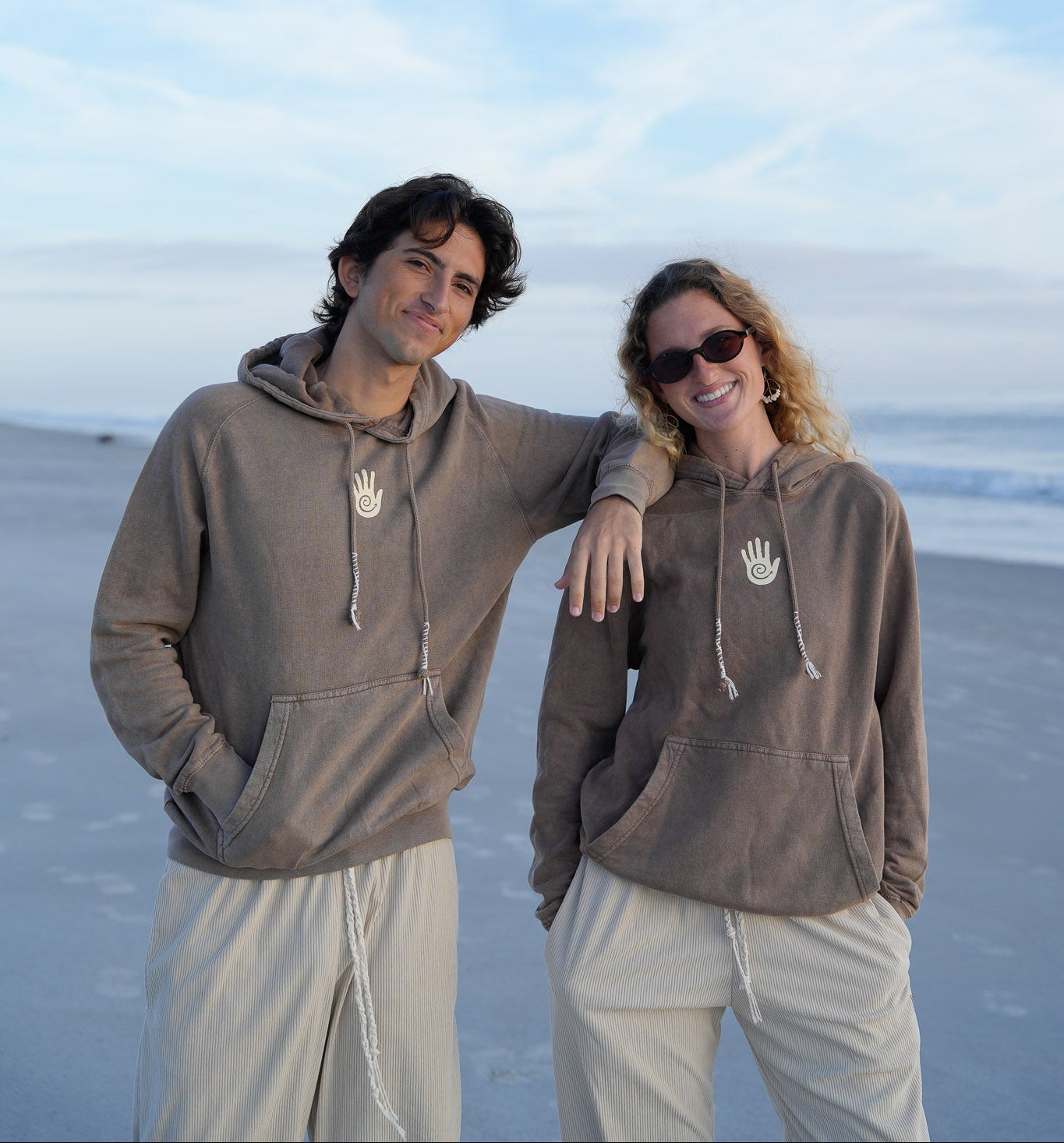 Two people wearing matching brown hoodies on a beach with a clear blue sky.