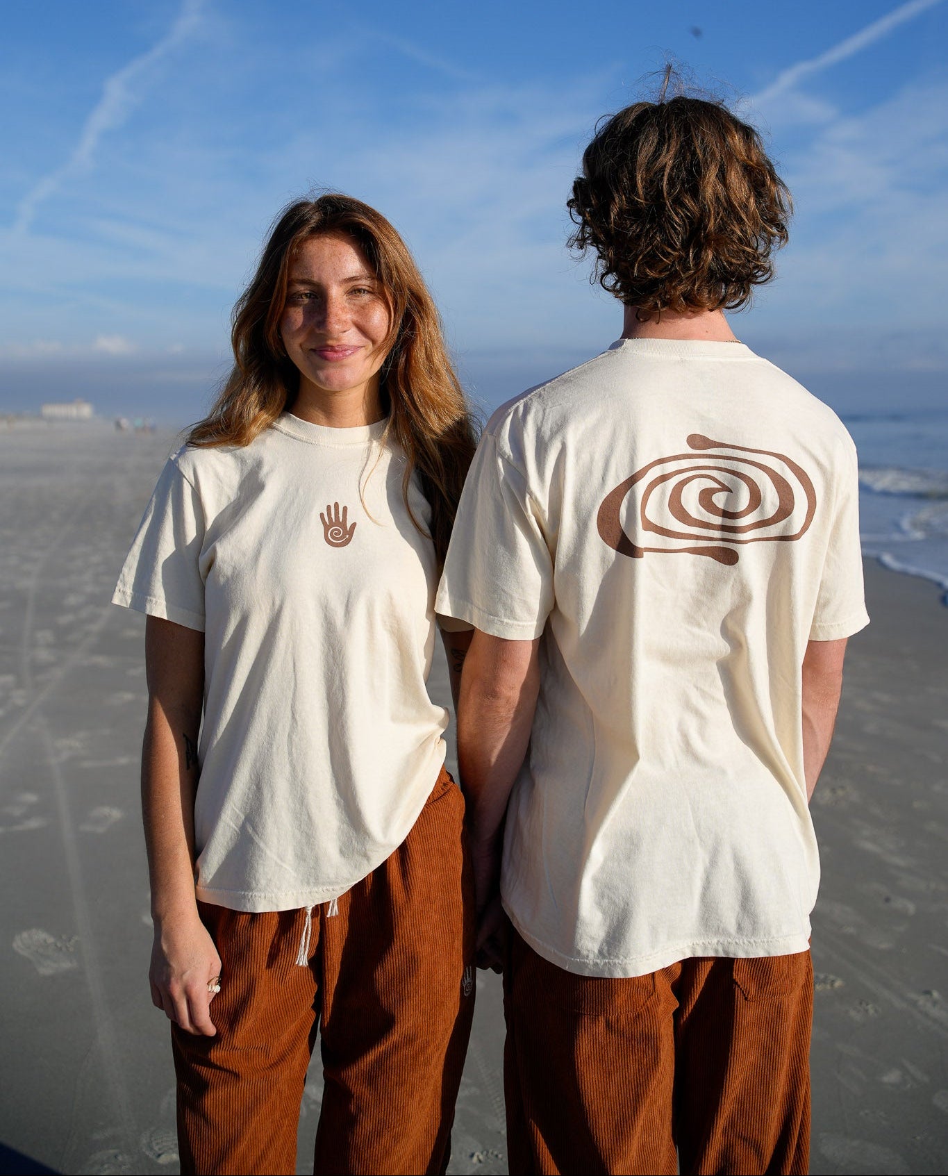 Two people standing on a beach wearing white t-shirts with hand and spiral designs.