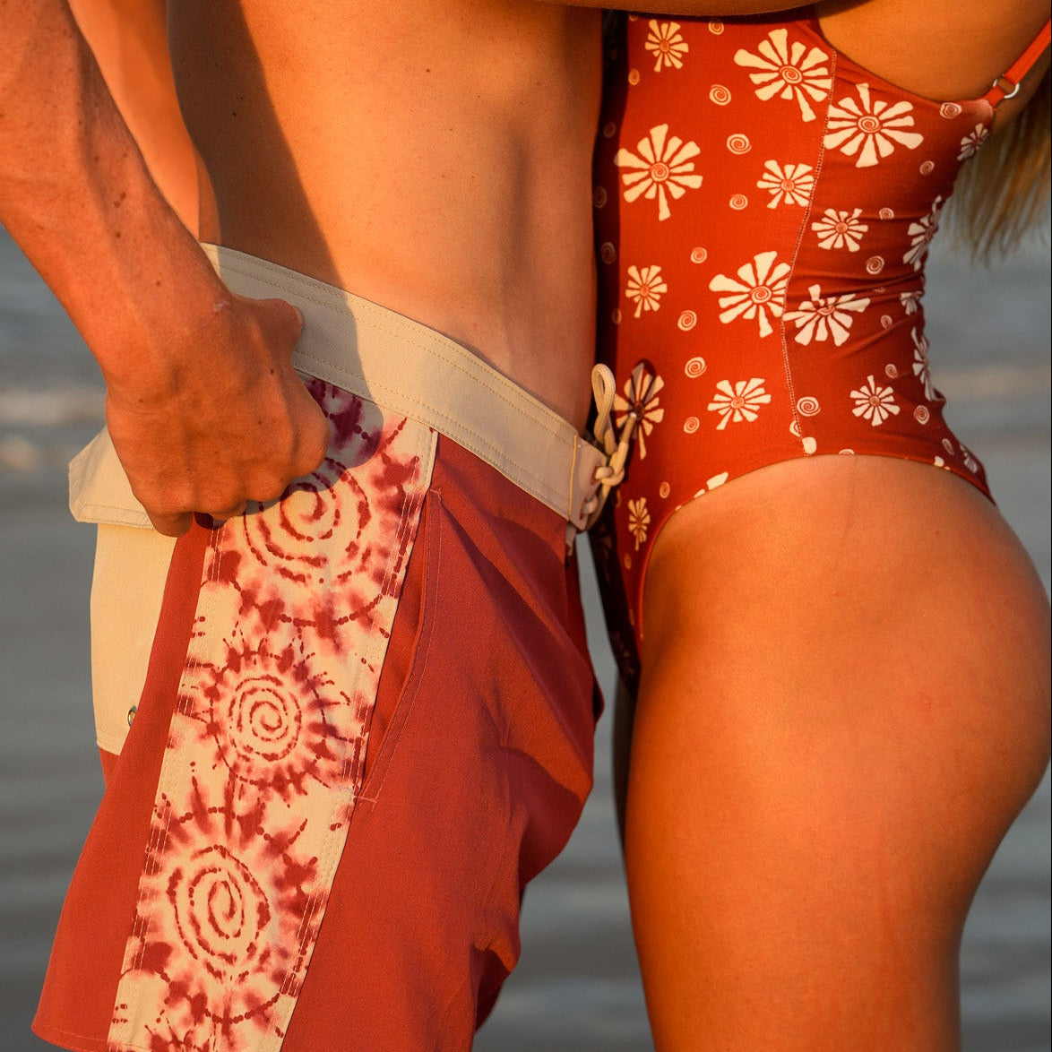 A couple embracing on a beach at sunset, with the man wearing rust-red and white patterned swim trunks and the woman wearing a matching rust-red one-piece swimsuit with white floral print.