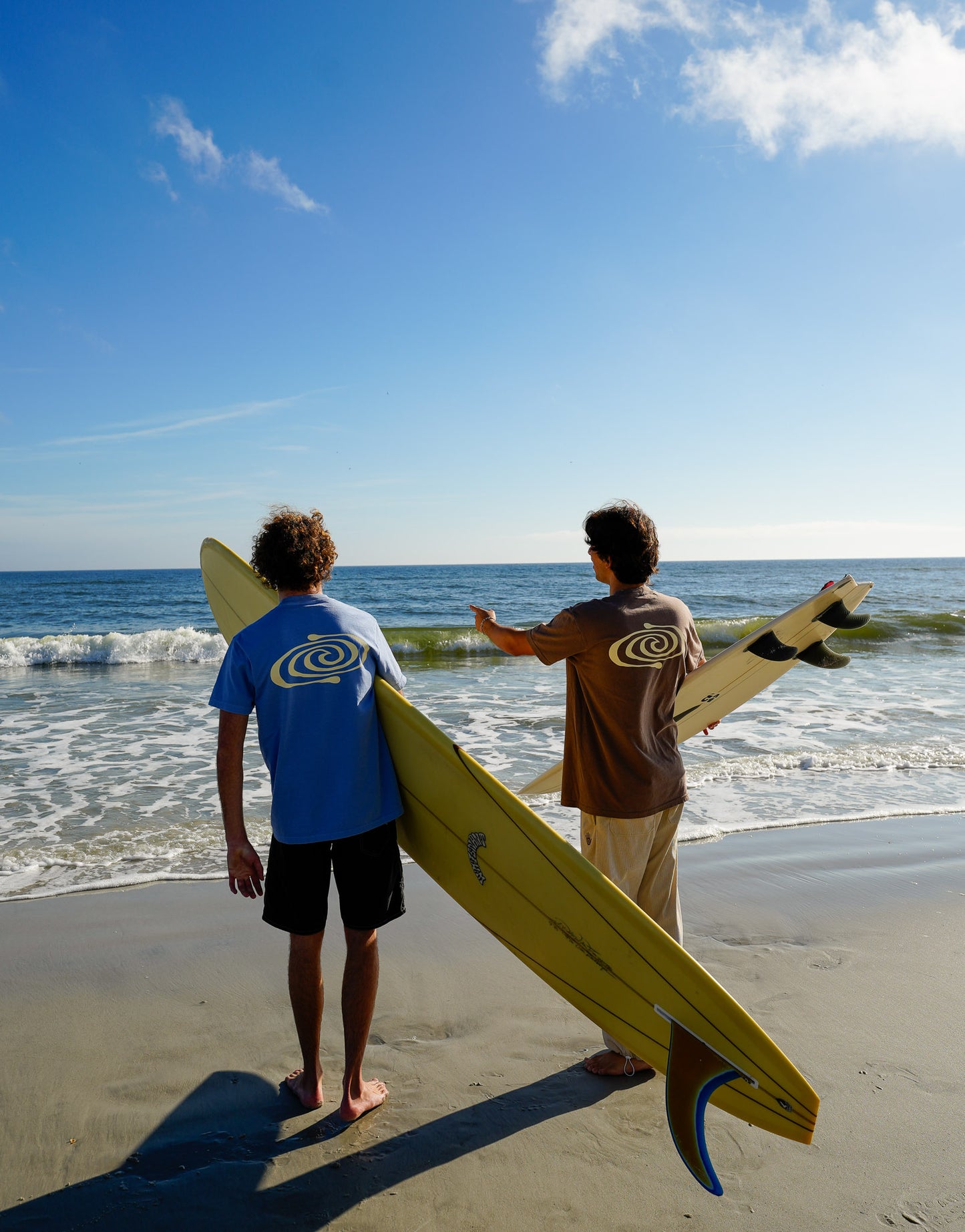 Two people holding surfboards on a beach with clear blue sky and ocean.