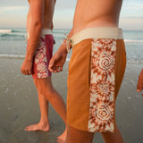 Men wearing patterned swim trunks on a beach at sunset, with one model in the foreground showing rust-orange shorts with a white tie-dye vertical panel and another model in the background wearing pink trunks.