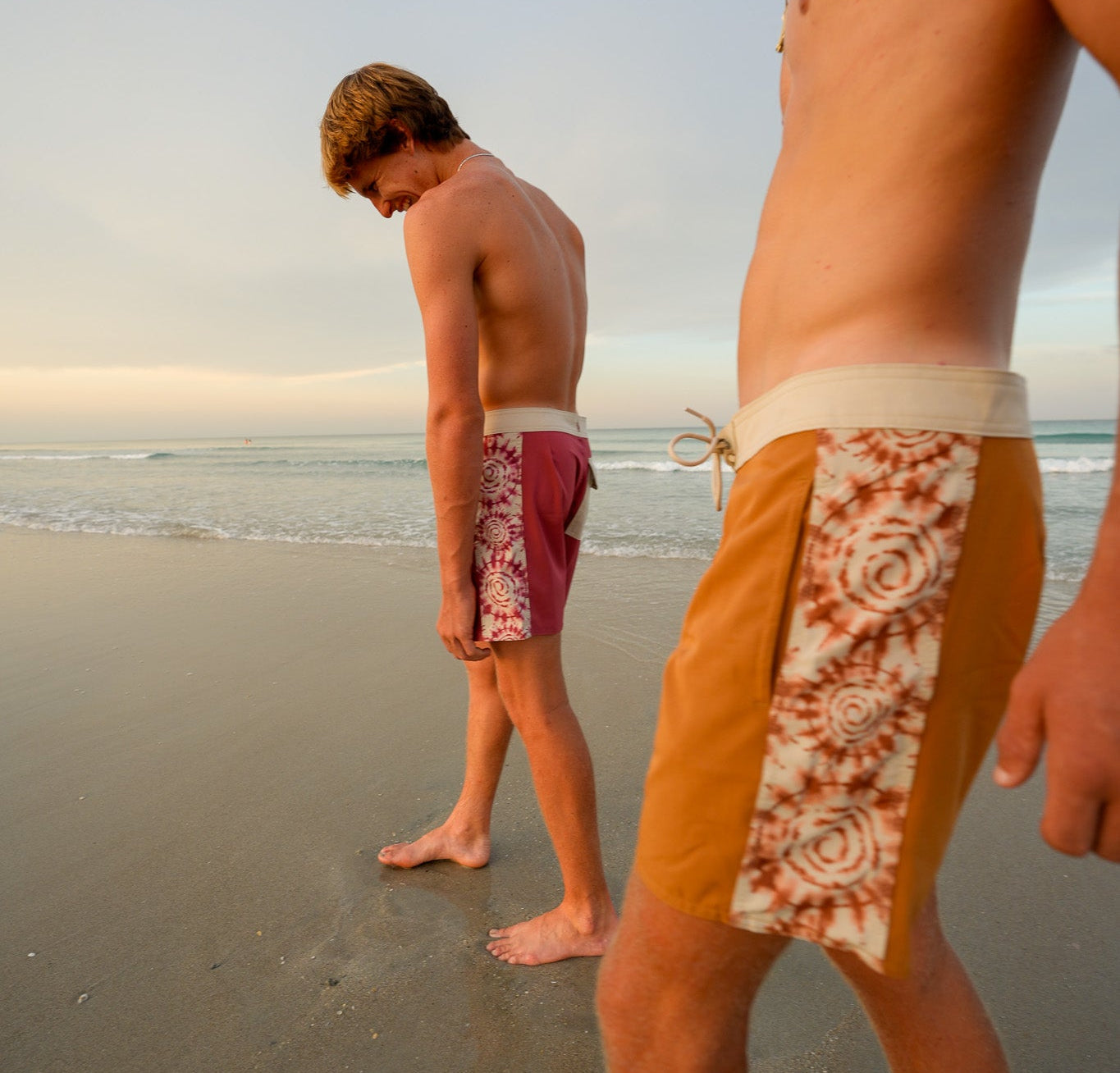 Two young men standing on a beach, wearing swim trunks. The man in the foreground is out of focus, while the man in the background is in sharper focus, looking down.