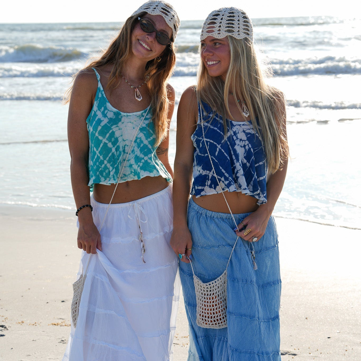 Two women standing on a sandy beach wearing matching bohemian-style two-piece sets with tie-dye crop tops and long flowing maxi skirts. One set is teal and white, and the other is blue and white, both paired with white crochet headwraps and crossbody bags.